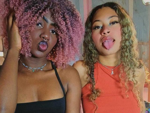 HaryAndKimmy with curly pink and blonde hair, sticking out tongues, wearing black and orange tops, black straps, nose ring, and necklace, in a playful, goofy pose.