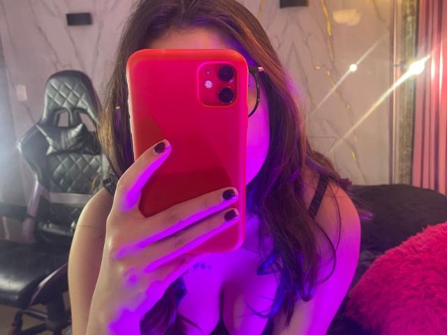 EmmaFeri with purple and pink lighting, holding a red phone covering her face, showing her large tits in a black bra, with long brown hair and black nails.