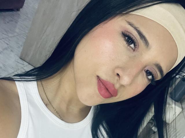 SophieCan has a cute face with black hair, pink lips, and a white headband. She's wearing a white tank top, lying on a bed, looking at the camera.