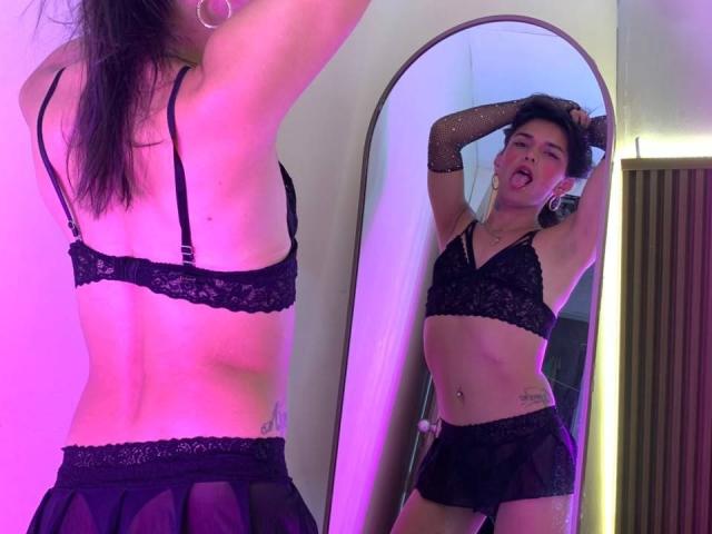 MaraSummers con un sujetador de encaje negro y shorts transparentes, vista de frente y de atrás en el espejo, sacando la lengua, con los brazos levantados, tatuaje visible, iluminación rosa y morada, cabello negro corto..