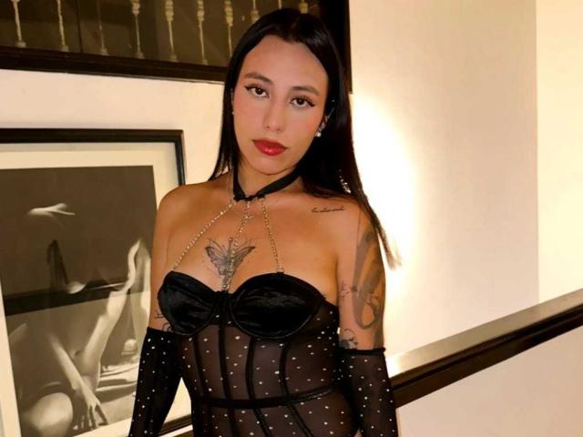 IvyLovely with black sheer polka-dot dress, strapless with black cups, chain collar, tattoos on chest and arms, standing in staircase, wearing red lipstick, looking at camera.