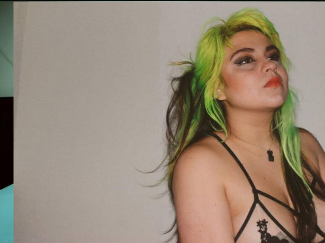 GineBralkino with neon green and black hair, wearing a black lace top, has pouty red lips, and a small black pendant, leaning against a white wall.