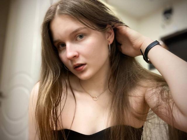 JuttaHillwig has long brown hair, pale skin, and small tits. She's wearing a black strap top, looking to the side with a slight open-mouth expression. Wearing a black wristband.