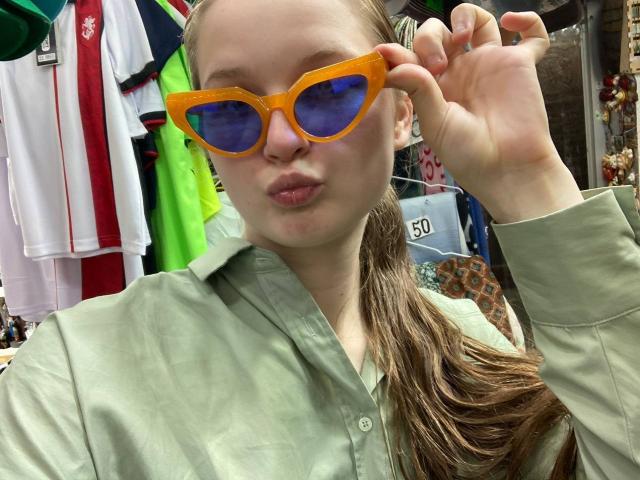 JuttaHillwig wearing bright orange sunglasses with blue lenses, pouting her lips, and holding the glasses with her right hand. She has light skin and brown hair in a ponytail.