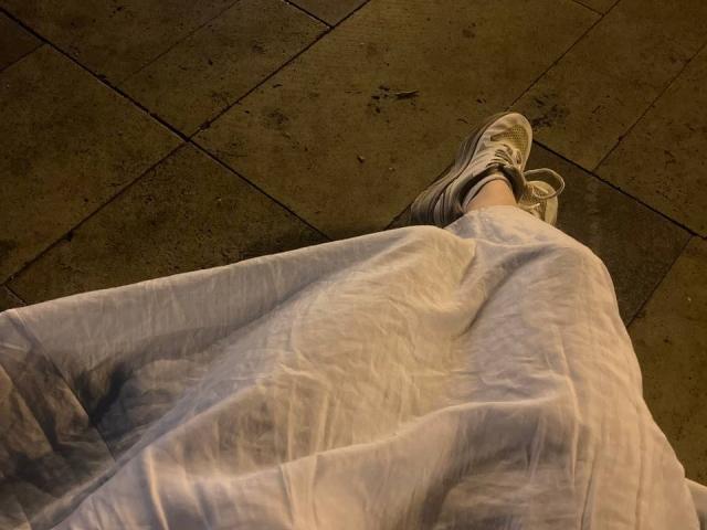 JuttaHillwig's legs in white crinkled pants, wearing gray and white sneakers, standing on brown tiled floor, with shadows on her pants.