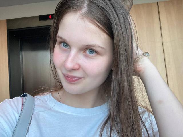 JuttaHillwig with blue eyes, fair skin, and brown hair, wearing a white shirt, has a slight smile, and is holding her hair up with her right hand.