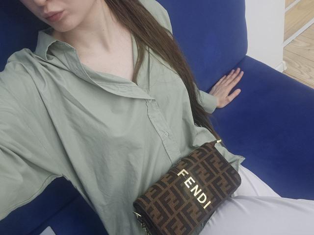 JuttaHillwig with long brown hair, green button-up shirt, white pants, blue sofa, and Fendi patterned purse, showing her tits slightly through shirt.