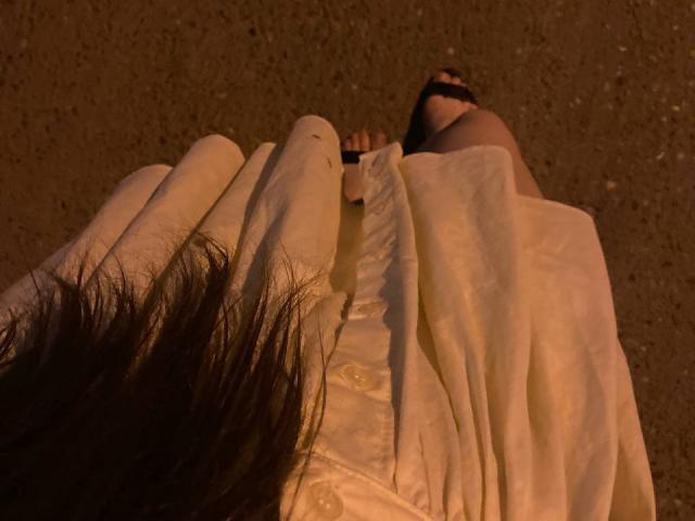 JuttaHillwig's hand holding white fabric over her bare pussy and tits, with her black-clad foot visible in the background, under dim, orange lighting.