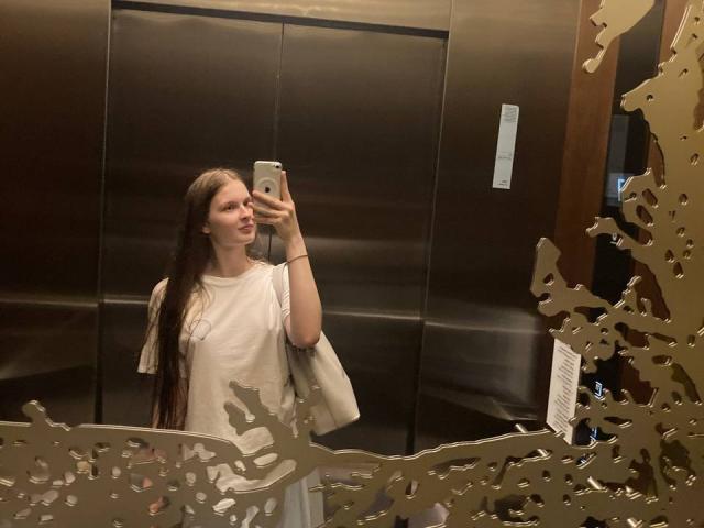 JuttaHillwig taking a selfie in a mirror, showing her long brown hair, white dress, and holding a smartphone with a subtle smile, standing in front of a reflective elevator.