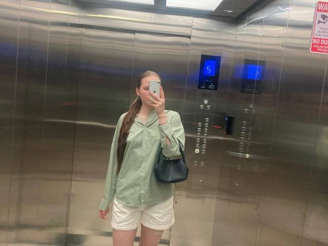 JuttaHillwig with long brown hair in a ponytail, wearing a green shirt and white shorts, holding a black bag, taking a mirror selfie in a metallic elevator.