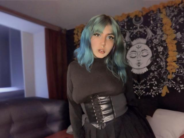 Alice Calloway, met haar turquoise blauwe haar, zwarte coltrui en korset, staat in een kamer en kijkt naar de camera, met een wandtapijt met een maanmotief achter haar. Haar borsten zijn zichtbaar..