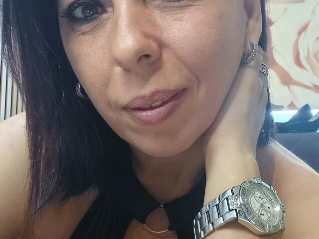 LauraSin with dark brown hair, winking, wearing a silver watch on her wrist, black necklace, and hoop earrings, smiling with her hand on her cheek, showing off her tits.