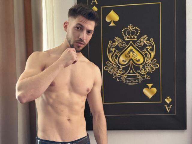 FelixRook, sem camisa, musculoso, com cabelo e barba curtos e escuros, segurando o queixo, em pé em frente a um cartão preto e dourado com um brasão real, vestindo calças pretas..