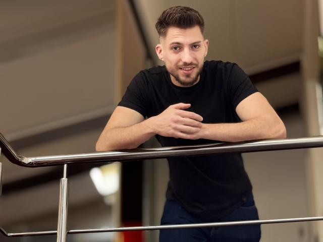 FelixRook, com cabelo castanho curto, camiseta preta e calças jeans azuis, apoiado em um corrimão de metal, mão direita no corrimão, mão esquerda no peito, sorrindo levemente..