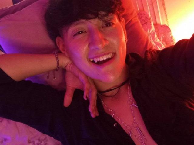 DannyRouds, con capelli ricci scuri, sorride a bocca aperta, indossa una maglietta nera e un collare, con una mano dietro la testa, illuminazione viola e rossa, denti visibili, braccialetto al polso..