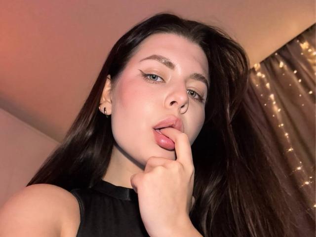 JoeyBickart with long brown hair, pale skin, and pink lips, sticking her finger into her mouth, wearing a black sleeveless top, looking seductive. Her tits are not visible.