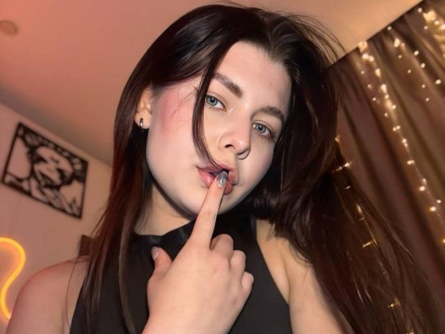 JoeyBickart with dark brown hair, pale skin, and blue eyes, sucking on her finger with a black sleeveless top, showing a cute, playful expression.