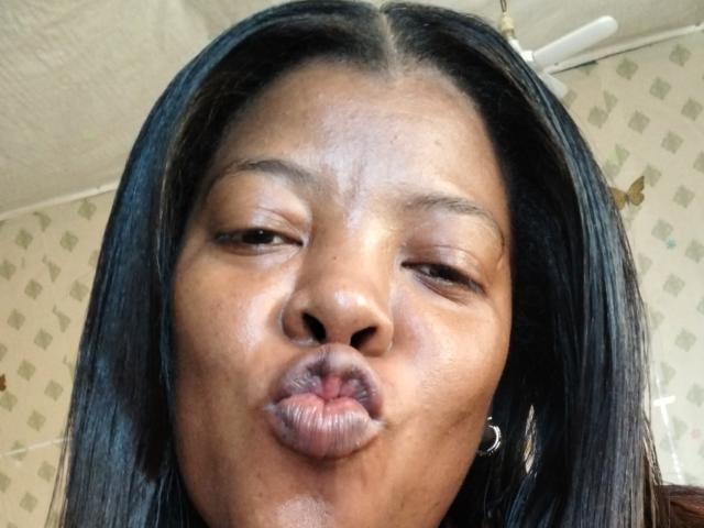 BrownieBabe's puffy ass-kissing face, with half-closed eyes and dark straight hair, wearing a small crescent moon earring. Her brown skin is shiny, and her lips are puckered.