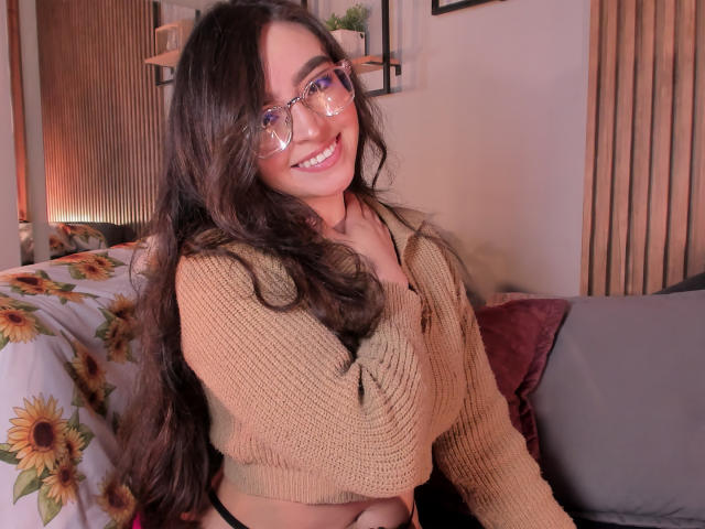 EvaMcAllister with long dark hair, glasses, smiling, wearing a tan knit top, showing her tits, sitting on a bed with sunflower-patterned sheets, black panties visible.