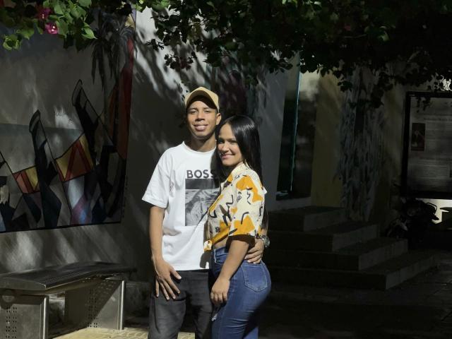 SofiaAndAndres with a tan boy, baseball cap, white shirt, black pants, standing next to a Latina girl, yellow pattern shirt, blue jeans, black hair, smiling, tree overhead.