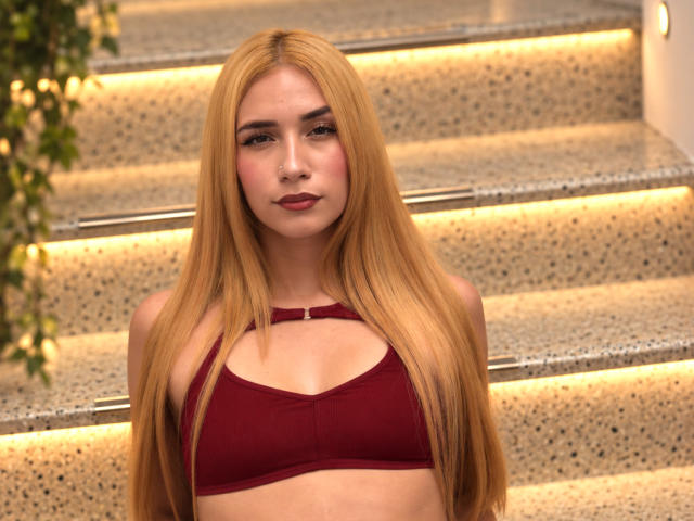 TanniaMoon has long blonde hair, a red bra, and a cute face, standing on illuminated stairs with a subtle smile, showing her small tits and smooth skin.