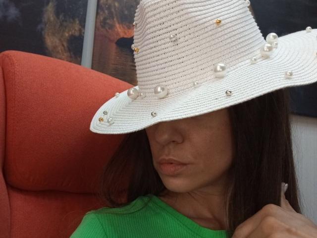 ViktoriiaLu, portant un chapeau de paille blanc orné de perles, une chemise verte, et faisant la moue, a les cheveux bruns et est assise sur une chaise orange..