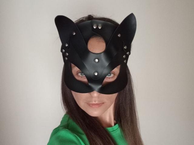 ViktoriiaLu porte un masque en cuir noir avec des oreilles de chat, des clous en métal et une découpe au niveau du nez, une chemise verte, de longs cheveux bruns et des yeux bleus, et elle regarde directement l'appareil photo..