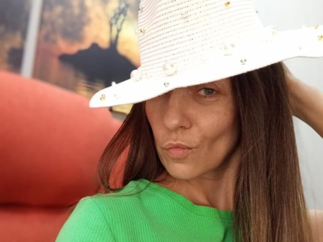 ViktoriiaLu lleva un sombrero blanco de paja con pequeñas cuentas, una camisa verde y frunce los labios. Su cabello castaño está suelto y está mirando a la cámara..