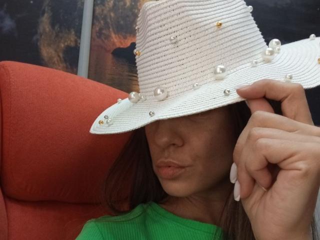 ViktoriiaLu portant un chapeau de soleil blanc texturé avec des ornements de perles, une chemise verte et des lèvres pulpeuses, tenant le chapeau avec sa main, montrant de longs ongles blancs..