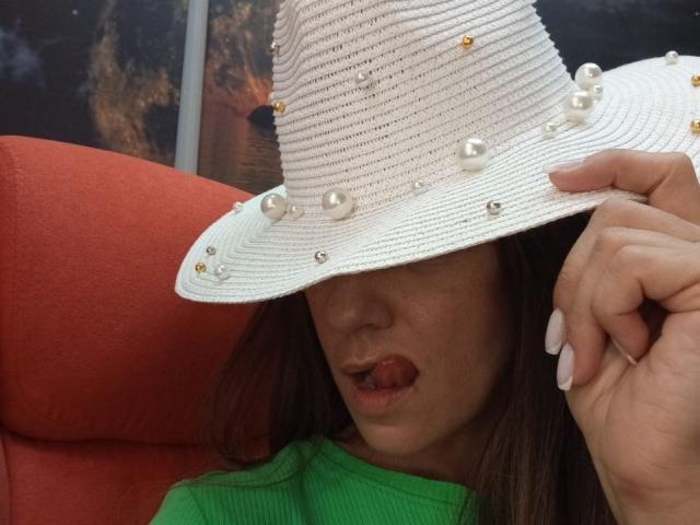 ViktoriiaLu, portant un chapeau de paille blanc orné de perles, la langue tirée, un t-shirt vert, des ongles longs, assise sur une chaise rouge, avec des cheveux bruns et une expression mignonne et espiègle..
