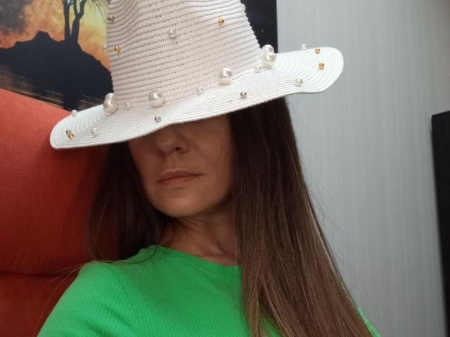 ViktoriiaLu, con cabello largo y castaño, lleva un sombrero de paja blanco con adornos de perlas y oro, y una camisa verde de punto acanalado que le cubre los ojos. En el fondo, hay una silla roja..