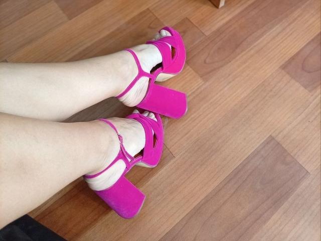 Les jambes pâles de ViktoriiaLu, chaussées de talons compensés rose vif avec de larges brides, se tiennent sur un plancher en bois. Ses pieds sont légèrement écartés, mettant en valeur ces chaussures éclatantes..