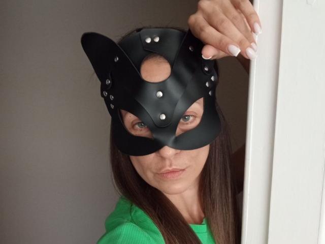 ViktoriiaLu with a black leather cat mask, showing her green eyes and pouty lips, has long brown hair and white nails, wearing a green hoodie, peeks from a doorway.