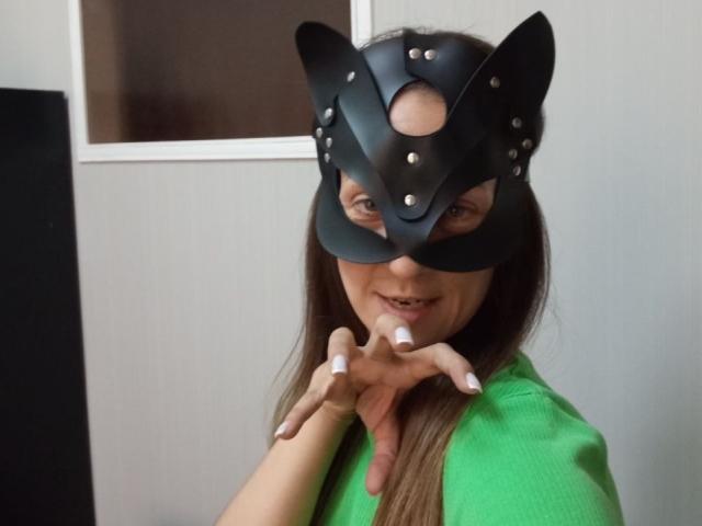 ViktoriiaLu portant un masque de chat en cuir noir avec des clous en métal, une chemise verte, de longs cheveux bruns, des ongles blancs, souriante, les mains près du visage. Les seins ne sont pas visibles. Les parties intimes ne sont pas visibles..