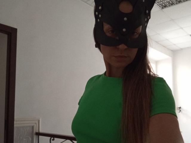 ViktoriiaLu indossa una maschera di gatto nera con dei fori, una maglietta a coste verde e lunghi capelli castani, mostrando i suoi piccoli seni, in una stanza poco illuminata..