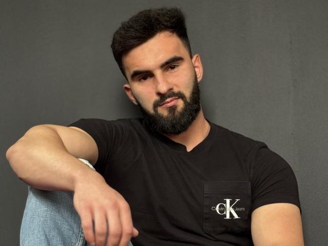MicahLane, een man met een baard en donker haar, gekleed in een zwart T-shirt, leunt op zijn knie, draagt jeans, met een grijze achtergrond, ontspannen, met een lichte glimlach, gemiddeld postuur, schattig, casual, zelfverzekerd, aantrekkelijk..