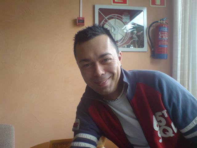 Dannyboy20-fun-hot, short-haired dude with a smile, wearing a red and blue jacket, leaning on a table, fire extinguisher on wall, brown wall, window on right.