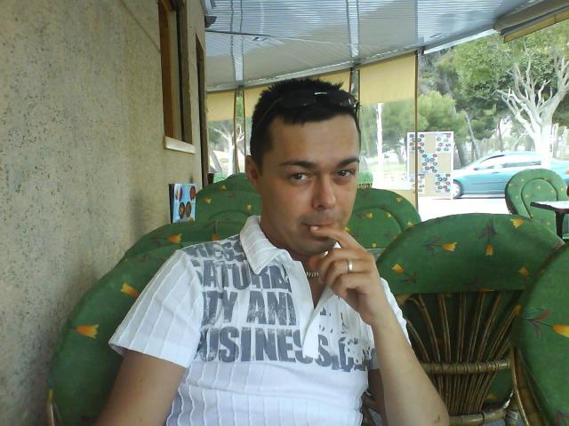 Dannyboy20-fun-hot with short black hair, light skin, and a white striped shirt, smoking a cigarette, sitting on a green, cactus-patterned chair, under a metal awning.