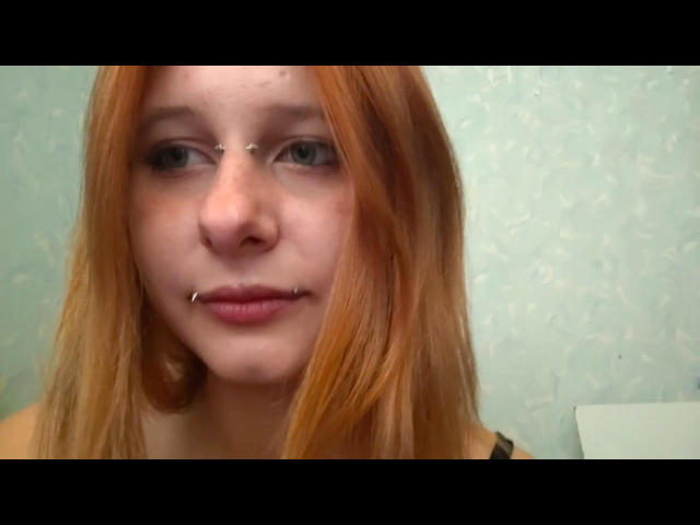 BasiliaGigger has red hair, a nose ring, and a lower lip piercing with a small metal bar. Her blue eyes are slightly open, and she's wearing a black top.