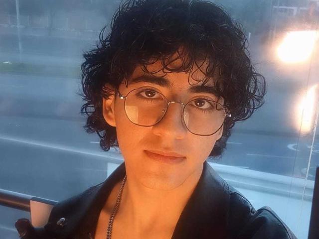 ElliotNoir with curly black hair, wearing round glasses and a black leather jacket, has a subtle smile and is looking at the camera in a selfie.