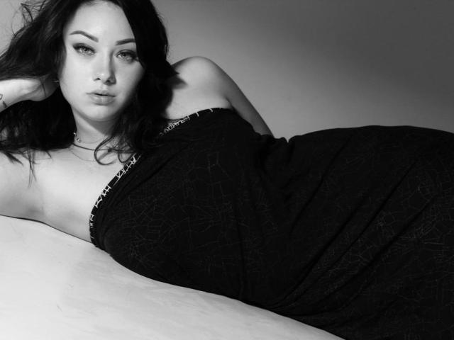 AdriannaGoldis with dark hair, lying on a bed, wearing a black, strapless dress, exposing her bare shoulders and upper chest, looking seductively at the camera, greyscale photo.