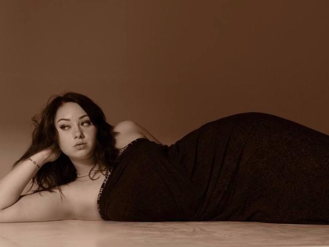AdriannaGoldis is lying on her side, dressed in a black, textured, strapless dress, with her hand under her head, and her ass prominently raised, looking at the camera.