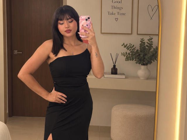 AshlyLee with black hair, wearing a black one-shoulder dress, showing her ass in a mirror selfie, holding a pink phone, standing in a room with a white vase.