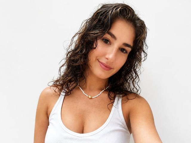AmandaKrous, com cabelo castanho escuro, úmido e encaracolado, blusa de algodão branca, seios pequenos, pele clara, sorriso discreto, colar e batom rosa claro em uma foto simples e clara..