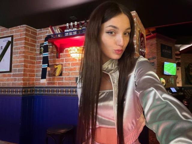 ElisVane with long brown hair, wearing a shiny pink and silver jacket, has a cute face and small tits, taking a selfie in a dimly lit bar.