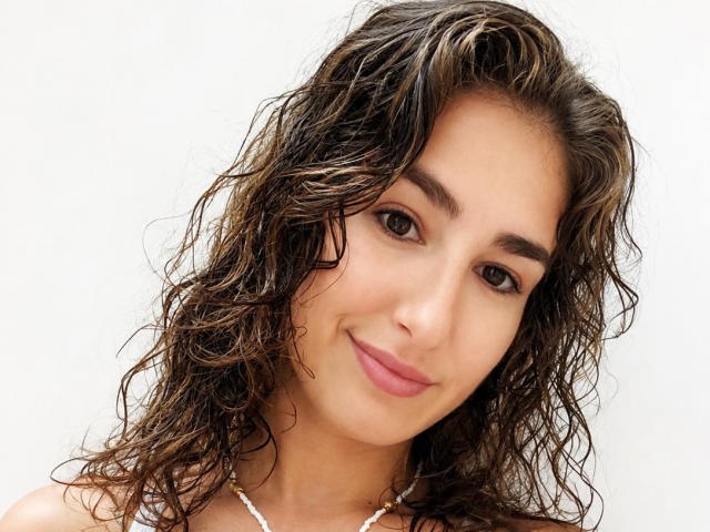 AmandaKrous has curly, wet brown hair, medium skin, and pink lips, smiling with brown eyes, wearing a white necklace, and looking at the camera.