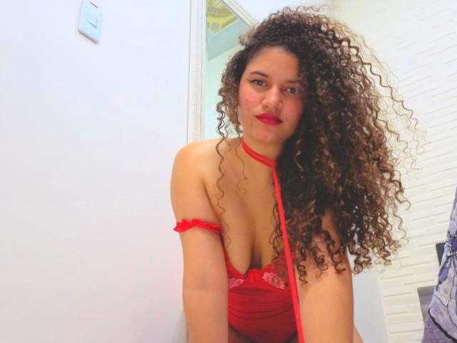 SanetPerlas with curly brown hair, red lingerie, and red strap, showing her tits, sitting in a white room, smiling with red lipstick.