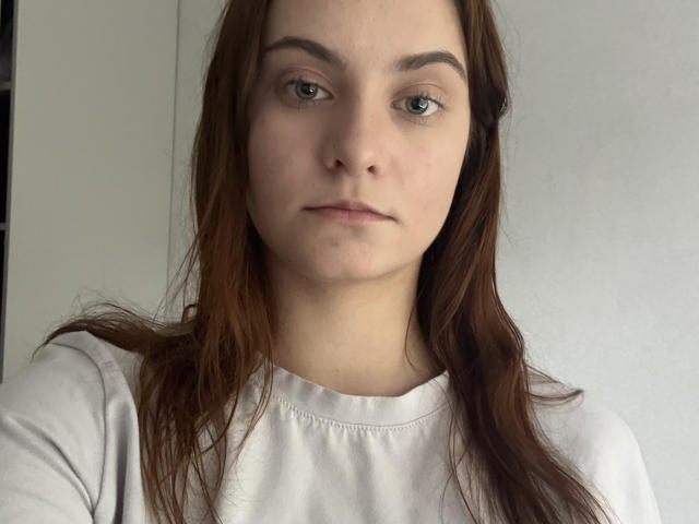 FriedaArnaldo has pale skin, green eyes, and brown wavy hair, wearing a white shirt, looking straight at the camera with a neutral expression.