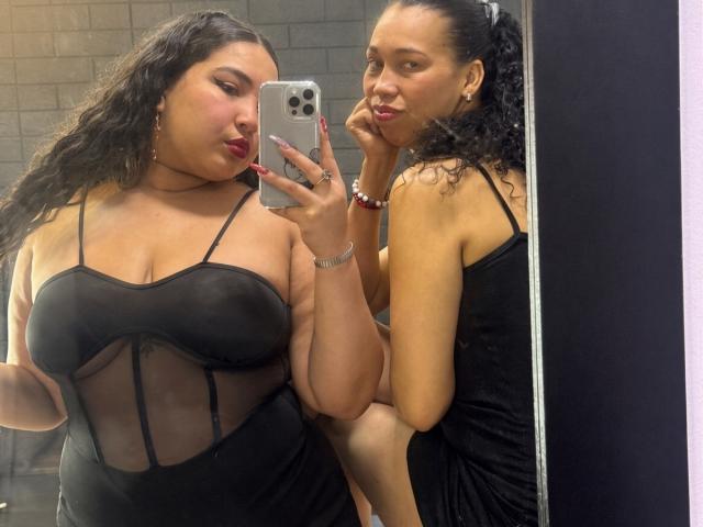 CharlotteTiffany with curly black hair, medium brown skin, and red lipstick, wearing a black sheer dress, showing her big boobs in a mirror selfie, pouting with her finger on her cheek.