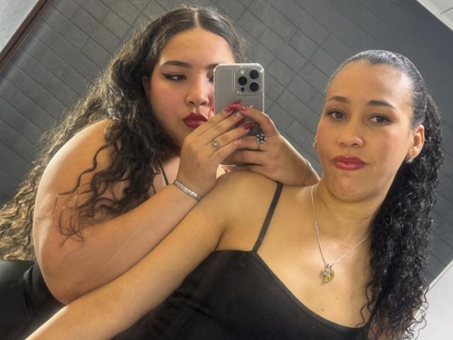 CharlotteTiffany with curly black hair, wearing a black tank top, holding a phone, standing behind CharlotteTiffany with black curly hair, wearing a black tank top, smiling.